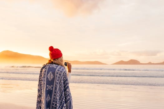 A woman wrapped in a blanket, wearing a red winter hat, enjoys a sunset at a serene beach with distant mountains.