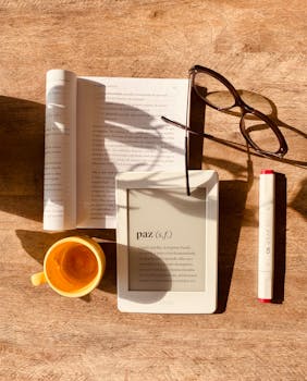 From above composition of e book and paper book on wooden table with cup and glasses in sunlight